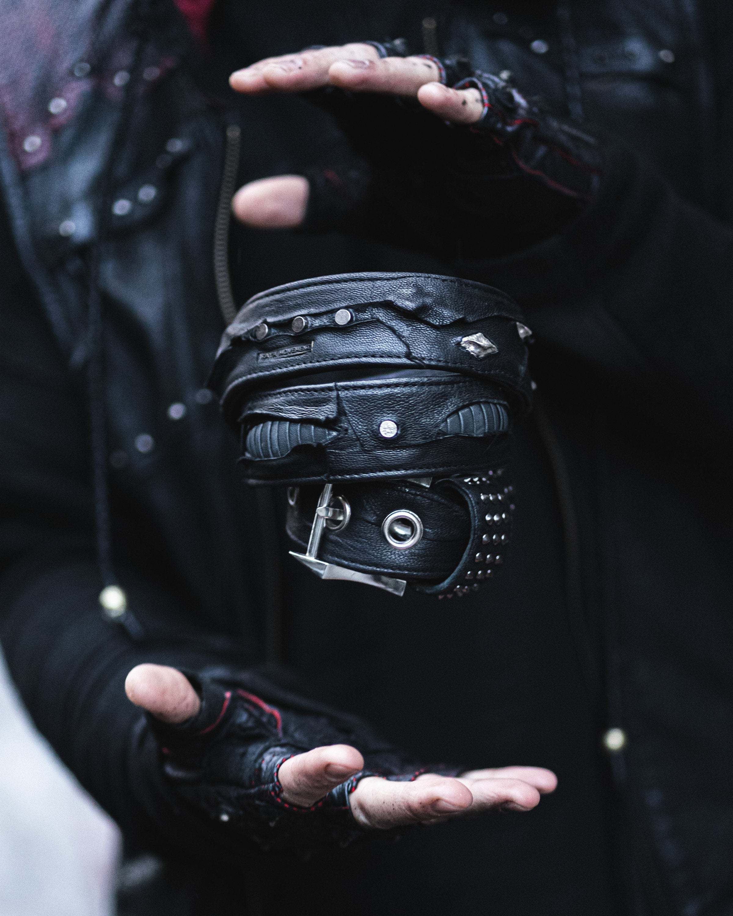 A person wearing fingerless gloves and a black leather outfit holds their hands around a levitating stack of KRAV Wasteland Men's Black Studded Leather Belts by Kaya Movement, adorned with silver spine studs and buckles, creating an illusion of the belts floating between their hands.